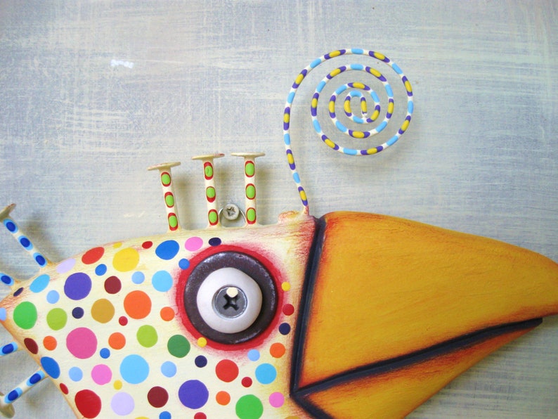 May include: A whimsical, colorful fish sculpture with a large yellow beak and a spiral accent. The fish's body is cream-colored with vibrant polka dots in various colors. It has a large, round eye and three small, decorative posts.