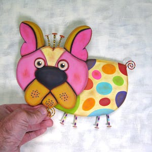 Puede incluir: Una figura de perro de madera colorida y caprichosa. El perro tiene una cara rosa, orejas amarillas y una nariz negra. Su cuerpo est&aacute; decorado con grandes lunares multicolores. Tiene peque&ntilde;as patas de metal y una cola rizada.