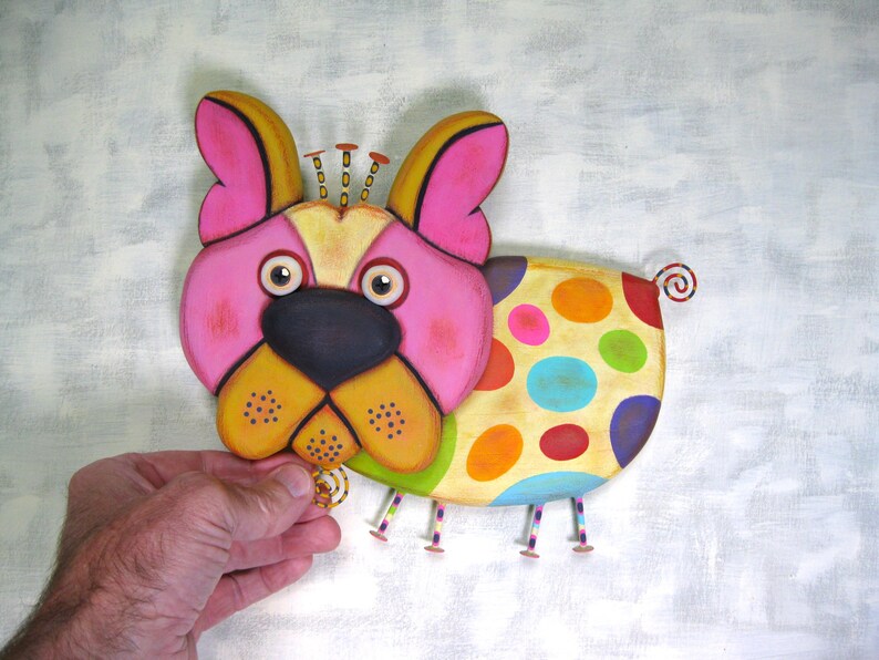 Puede incluir: Una figura de perro de madera colorida y caprichosa. El perro tiene una cara rosa, orejas amarillas y una nariz negra. Su cuerpo est&aacute; decorado con grandes lunares multicolores. Tiene peque&ntilde;as patas de metal y una cola rizada.