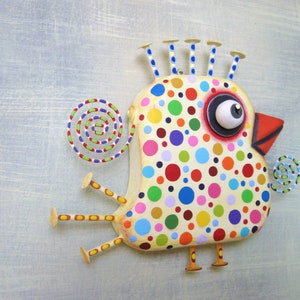 May include: A whimsical, handcrafted bird sculpture with a cream-colored body adorned with colorful polka dots. The bird features a large, expressive eye, a red beak, and decorative elements like a spiral tail and spiky head crests. The legs and feet are also visible.