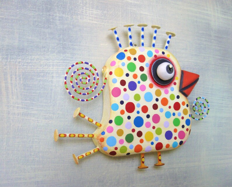 May include: A whimsical, handcrafted bird sculpture with a cream-colored body adorned with colorful polka dots. The bird features a large, expressive eye, a red beak, and decorative elements like a spiral tail and spiky head crests. The legs and feet are also visible.