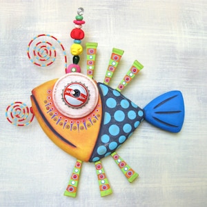 May include: A whimsical, handcrafted wooden fish sculpture. The fish features a vibrant color palette of yellow, blue, orange, and green. It has a bottle cap eye and is adorned with decorative elements like polka dots, spirals, and beaded accents. The sculpture is designed for wall hanging.