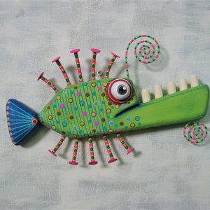 May include: A whimsical, colorful wooden fish sculpture. The fish features a green body with pink, blue, and yellow polka dots and stripes. It has a large, expressive eye, a green mouth with white teeth, and pink-tipped appendages. The tail is blue with a fan-like design.