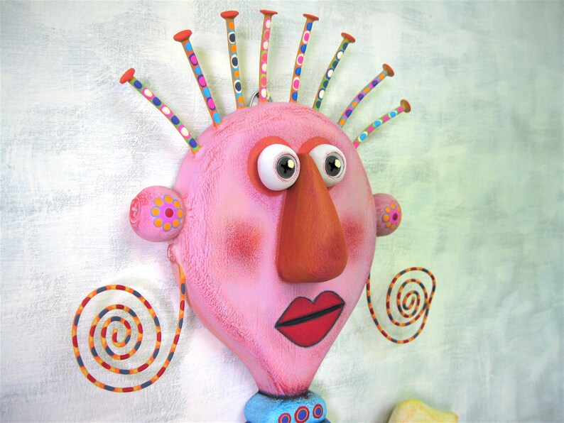 May include: A pink, whimsical face with large eyes, a long orange nose, and red lips. The face has a colourful, spiky hairdo and spiral-shaped earrings.