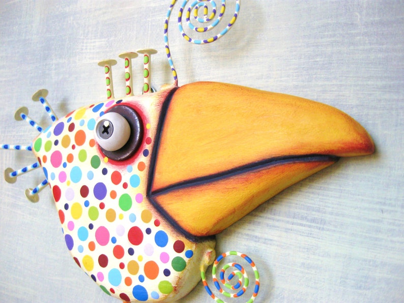 May include: A whimsical, colorful bird sculpture with a large yellow beak and a body covered in polka dots. The bird has a large, round eye and decorative metal accents. The sculpture is painted in bright, cheerful colors, making it a fun piece of art.