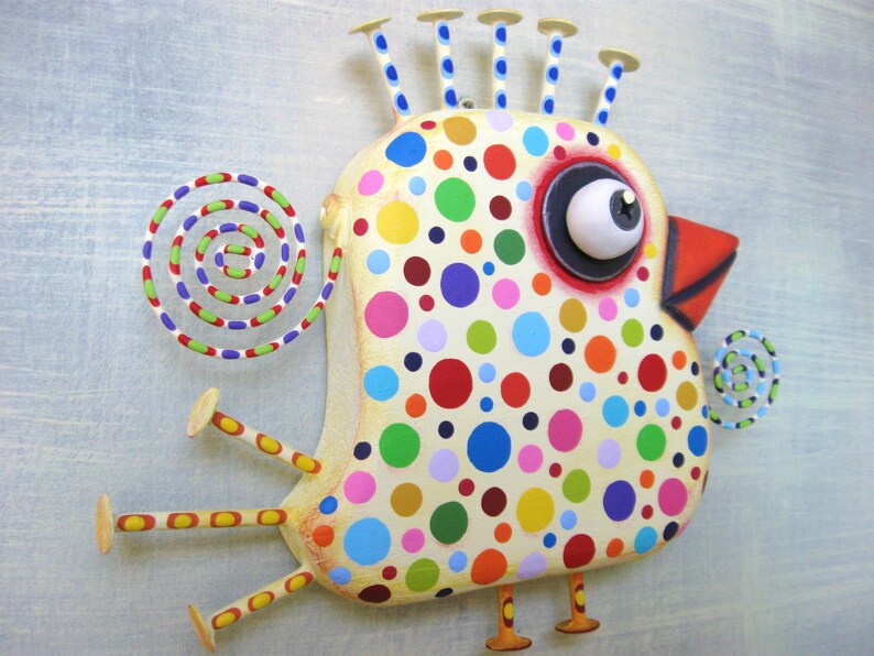 May include: A whimsical, colorful bird sculpture with a cream-colored body covered in multi-colored polka dots. It has a large eye, a red beak, and decorative spirals. The bird has stick-like legs and a crown of small posts.