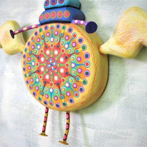 May include: A whimsical wooden sculpture of a bird with yellow wings, a blue and yellow body, and a colourful mandala design. The bird has long, thin legs with pink and orange stripes.