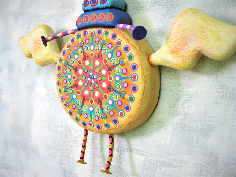 May include: A whimsical wooden sculpture of a bird with yellow wings, a blue and yellow body, and a colourful mandala design. The bird has long, thin legs with pink and orange stripes.