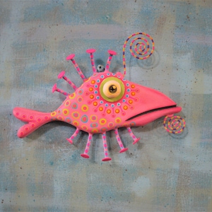 May include: A whimsical, pink fish sculpture with a large, central eye and colorful dot patterns. It features multiple pink appendages and a spiral detail, mounted on a textured, blue-green background.