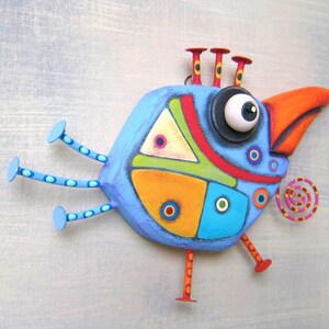 May include: A whimsical, hand-painted bird sculpture in shades of blue, orange, green, and red. The bird features a large eye, a curved orange beak, and decorative circles. It has three legs and three antenna-like structures on its head.