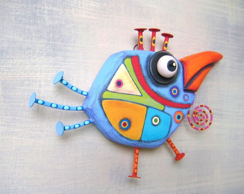 May include: A whimsical, hand-painted bird sculpture in shades of blue, orange, green, and red. The bird features a large eye, a curved orange beak, and decorative circles. It has three legs and three antenna-like structures on its head.