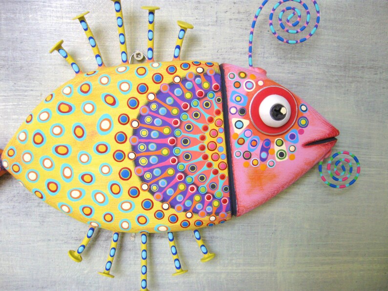 Puede incluir: Escultura de pez pintada a mano con una paleta de colores vibrantes. El pez presenta un cuerpo amarillo con lunares azules, naranjas y blancos. La cabeza es rosa con un ojo grande y expresivo. Clavos decorativos y un espiral a&ntilde;aden al dise&ntilde;o &uacute;nico.