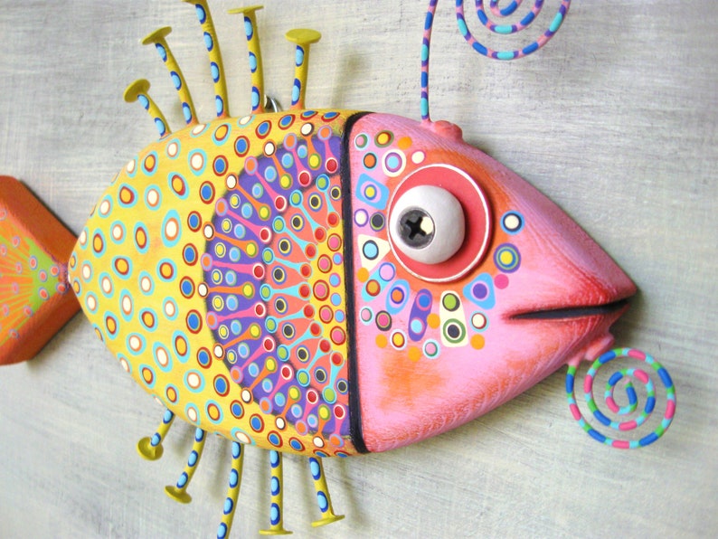 Puede incluir: Una colorida y caprichosa escultura de pez con cuerpo amarillo y rosa. El pez presenta c&iacute;rculos y patrones decorativos en varios colores, detalles met&aacute;licos y un detalle en espiral. La escultura est&aacute; montada sobre una superficie gris claro.