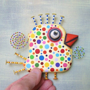 May include: A whimsical, hand-painted bird sculpture with a cream-colored body covered in colorful polka dots. It features a large, expressive eye, a red beak, and stylized legs and head accents. The artwork includes spiral accents on a light blue background.