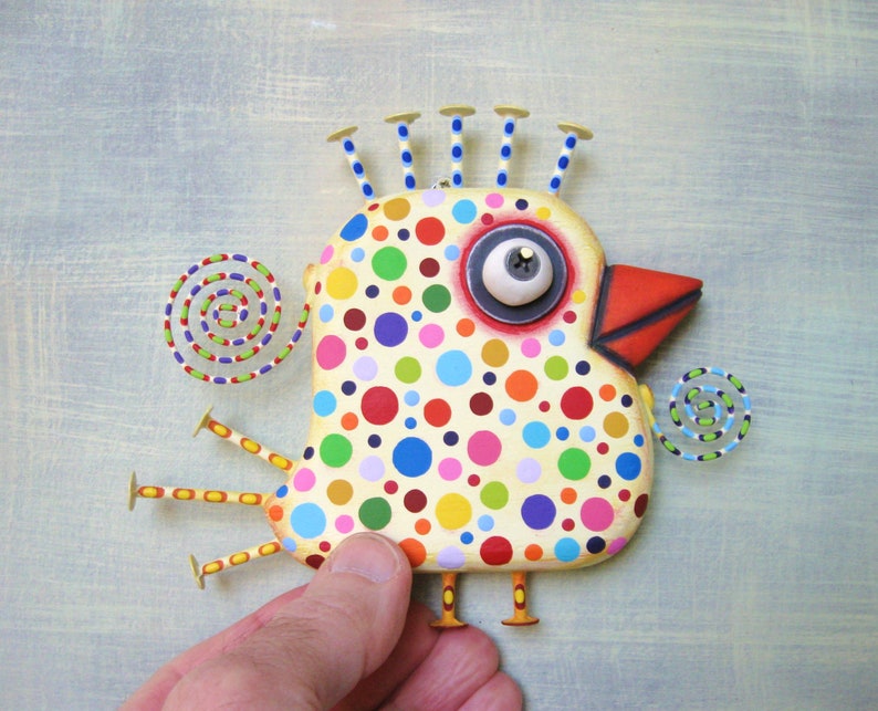 May include: A whimsical, hand-painted bird sculpture with a cream-colored body covered in colorful polka dots. It features a large, expressive eye, a red beak, and stylized legs and head accents. The artwork includes spiral accents on a light blue background.