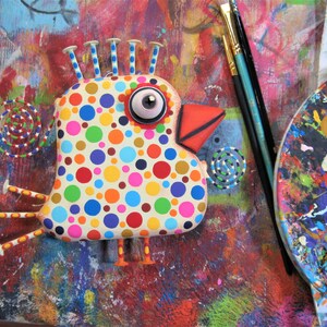 May include: A whimsical, colorful bird sculpture with a polka-dot pattern in various colors. The bird has a large eye, a red beak, and decorative accents. Paintbrushes and a palette are in the background, suggesting an art theme.