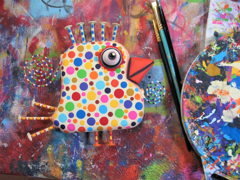 May include: A whimsical, colorful bird sculpture with a polka-dot pattern in various colors. The bird has a large eye, a red beak, and decorative accents. Paintbrushes and a palette are in the background, suggesting an art theme.
