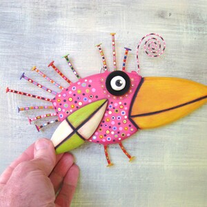 May include: A whimsical pink and orange bird sculpture with a large beak, black button eyes, and a green wing. The bird is decorated with colorful polka dots and has a spiral antenna on top of its head.