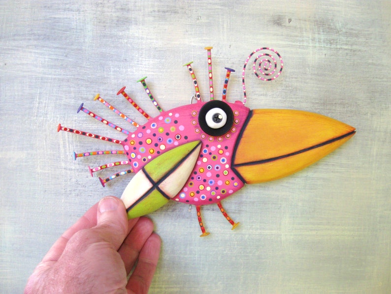 May include: A whimsical pink and orange bird sculpture with a large beak, black button eyes, and a green wing. The bird is decorated with colorful polka dots and has a spiral antenna on top of its head.