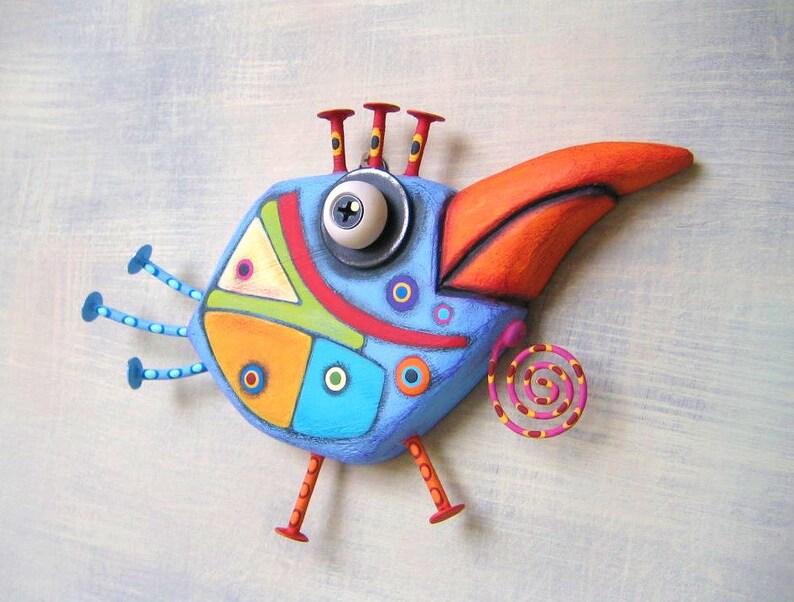 May include: A whimsical, colorful bird sculpture with a blue body, orange beak, and red accents. The bird has a large eye, three antennae, and decorative legs. The design incorporates geometric shapes and a spiral tail, creating a playful, artistic aesthetic.