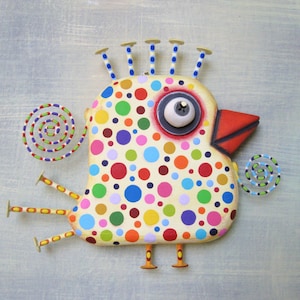 May include: A whimsical, handcrafted bird sculpture with a cream-colored body covered in colorful polka dots. It features a red beak, a large eye, and decorative accents, including spiral shapes and nail-like protrusions. The bird is mounted on a light blue background.