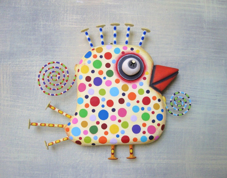 May include: A whimsical, handcrafted bird sculpture with a cream-colored body covered in colorful polka dots. It features a red beak, a large eye, and decorative accents, including spiral shapes and nail-like protrusions. The bird is mounted on a light blue background.