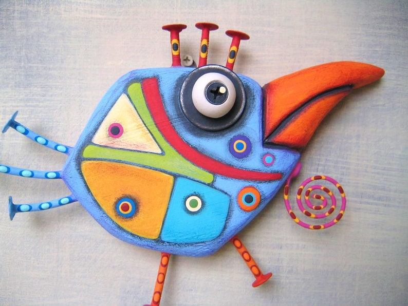 May include: A whimsical, colorful bird sculpture with a blue body, orange beak, and red accents. The bird has a large eye, decorative legs, and a spiral tail. The artwork is a unique, handcrafted piece.