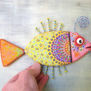 Puede incluir: Una escultura de pez de madera pintada a mano. El pez presenta un cuerpo amarillo con lunares azules y blancos, una cabeza rosa y una cola naranja. Tiene detalles decorativos de clavos y un detalle en espiral, creando una est&eacute;tica de arte popular vibrante.