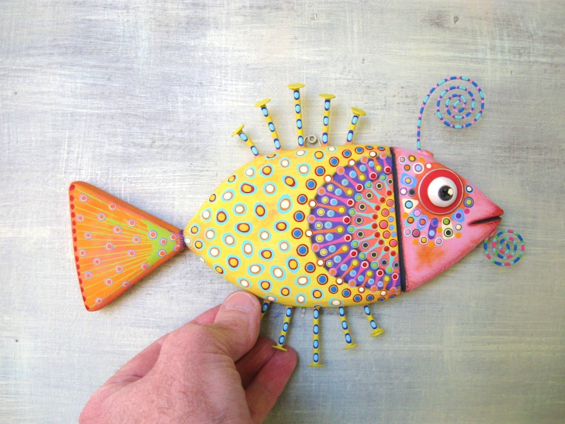 Puede incluir: Una escultura de pez de madera pintada a mano. El pez presenta un cuerpo amarillo con lunares azules y blancos, una cabeza rosa y una cola naranja. Tiene detalles decorativos de clavos y un detalle en espiral, creando una est&eacute;tica de arte popular vibrante.