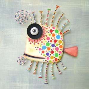 May include: A whimsical, colorful fish sculpture with a large, black eye and a white pupil. The fish is covered in polka dots of various colors, including red, blue, green, yellow, and purple. The fish has a long, thin tail and many thin, colorful legs.