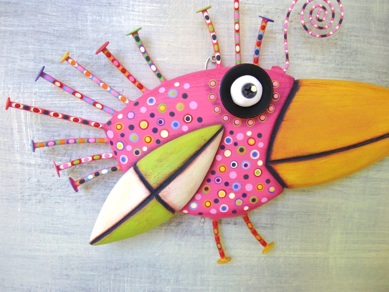 May include: A whimsical pink bird with a long orange beak, green and white wings, and a black button eye. The bird has a colorful, polka-dotted body and a spiral antenna on its head.