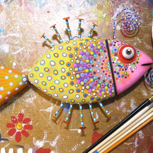 Puede incluir: Escultura de pez de madera pintada a mano. El pez tiene un cuerpo amarillo con c&iacute;rculos azules y blancos, una cabeza rosa y una cola naranja. Tiene aletas decorativas en forma de clavo y un ojo grande y expresivo. La obra de arte est&aacute; sobre una superficie salpicada de pintura.