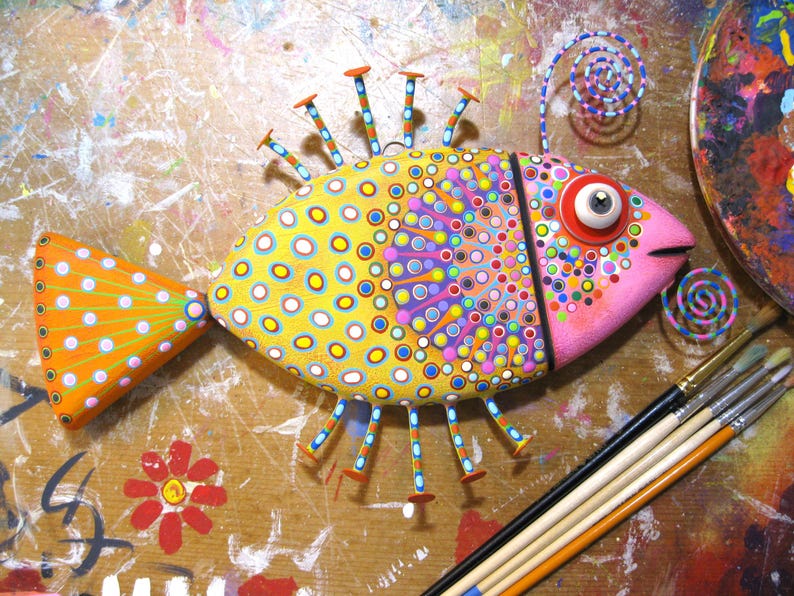 Puede incluir: Escultura de pez de madera pintada a mano. El pez tiene un cuerpo amarillo con c&iacute;rculos azules y blancos, una cabeza rosa y una cola naranja. Tiene aletas decorativas en forma de clavo y un ojo grande y expresivo. La obra de arte est&aacute; sobre una superficie salpicada de pintura.