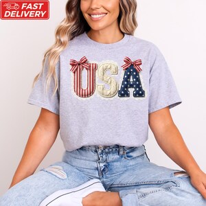 A woman wearing a grey t-shirt with the word "USA" embroidered on it, sitting on the floor with her legs crossed.