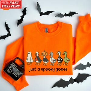 a bright orange sweatshirt with a Halloween-themed design, including a skeleton, jack-o-lantern, and a goose, along with a black mug with the text "Just a spooky goose". The mug is placed next to the sweatshirt, which is surrounded by black bats.