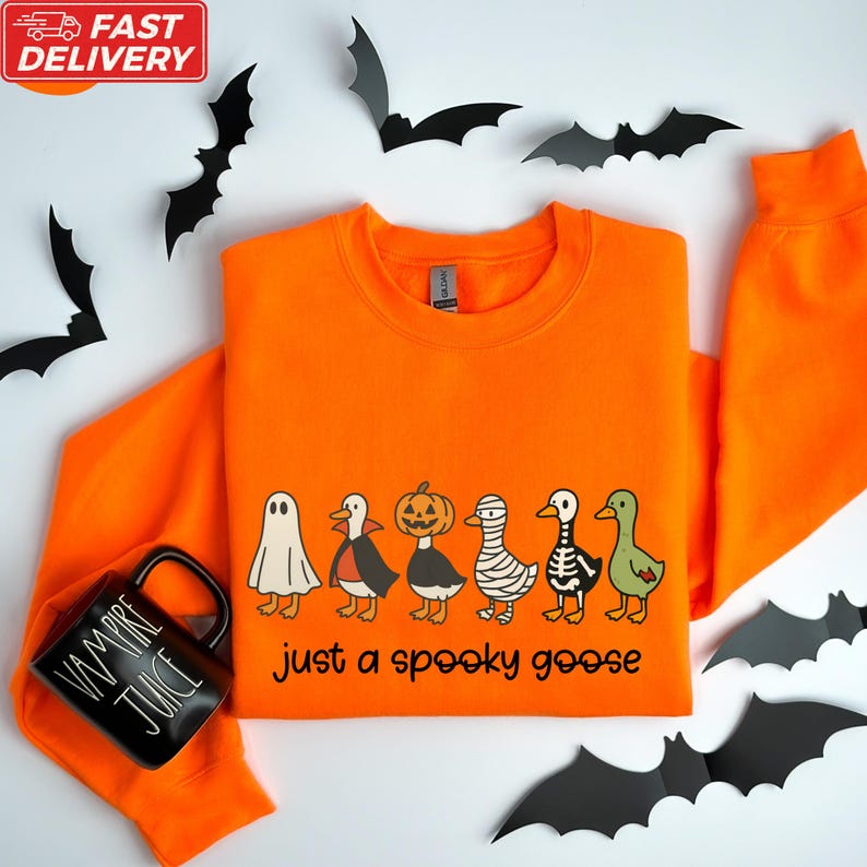 a bright orange sweatshirt with a Halloween-themed design, including a skeleton, jack-o-lantern, and a goose, along with a black mug with the text "Just a spooky goose". The mug is placed next to the sweatshirt, which is surrounded by black bats.