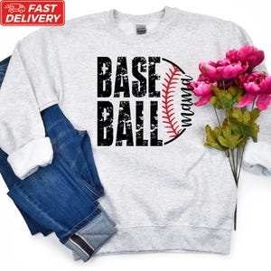A gray sweatshirt with the text "BASE BALL" printed on it, along with a baseball graphic and the word "Momma" below it. The sweatshirt is placed next to a pair of blue jeans and a bouquet of pink flowers.