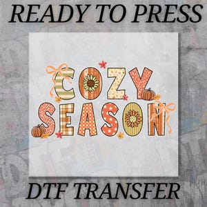 May include: A DTF transfer design with the words "COZY SEASON" in colorful, patterned letters. The design includes sunflowers, pumpkins, and fall leaves. The text "READY TO PRESS" and "DTF TRANSFER" are above and below the main text.