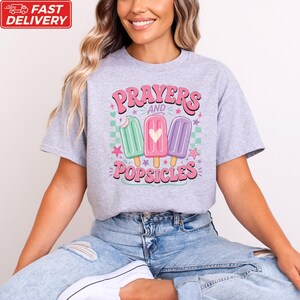 A woman wearing a gray t-shirt with a graphic design of three popsicles and the text "Prayers and Popsicles" printed on it.