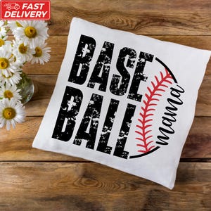 A white pillow with the text "BASE BALL" printed on it, along with a red baseball graphic, is placed on a wooden surface next to a vase of white flowers.