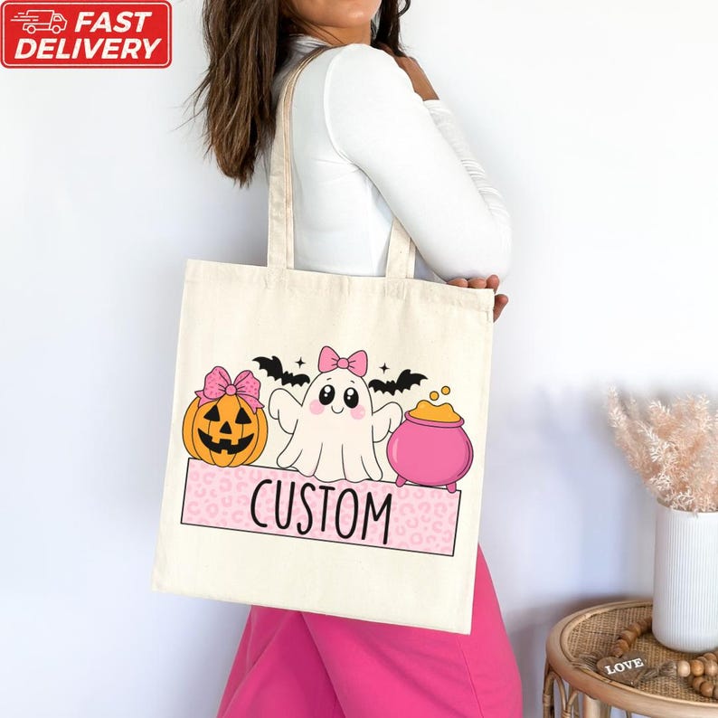 A woman is holding a white tote bag with a Halloween-themed design, including a ghost, pumpkin, and a pink bow.