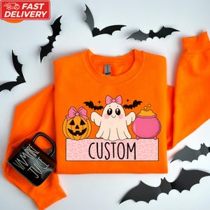 a bright orange sweatshirt with a custom design, including a pumpkin, ghost, and a pink pot, along with a black mug with the word "VAMPIRE JUICE" printed on it. The sweatshirt is surrounded by black bats, and the word "CUSTOM" is displayed on the sweatshirt.