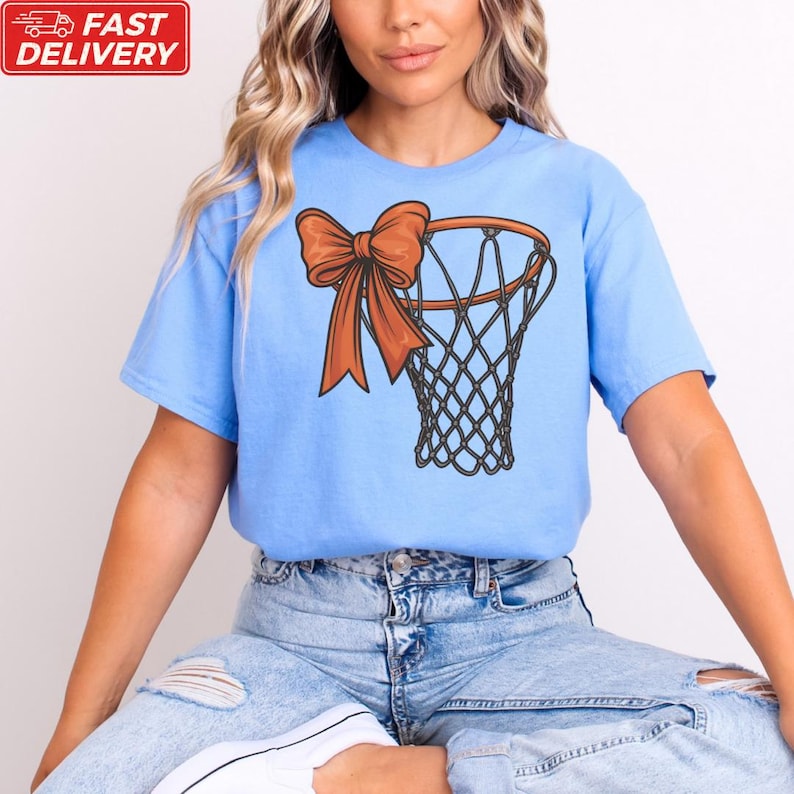 A woman wearing a blue t-shirt with a graphic of a basketball hoop and a bow on it, sitting on the ground.