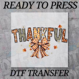 May include: A DTF transfer design with the word "THANKFUL" in a fall-themed font. The letters have plaid, leopard, and floral patterns. A large bow with a leopard print design is at the bottom, surrounded by leaves and daisies. The text "READY TO PRESS" and "DTF TRANSFER" are at the top and bottom.