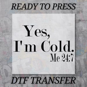 May include: A white square DTF transfer with the text "Yes, I'm Cold. Me 24:7" in black. The words "READY TO PRESS" and "DTF TRANSFER" are above and below the square. The background is a gray, textured surface.