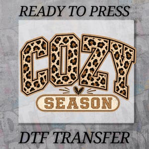 May include: A DTF transfer sheet featuring the text READY TO PRESS at the top and DTF TRANSFER at the bottom. The central graphic displays the word COZY in a large block font with a brown and black leopard print pattern, above the word SEASON.
