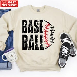 A beige sweatshirt with the text "BASE BALL" printed on it, along with a baseball graphic and a red baseball glove. Next to the sweatshirt are a pair of black and white sneakers, and a white mug with a floral design.