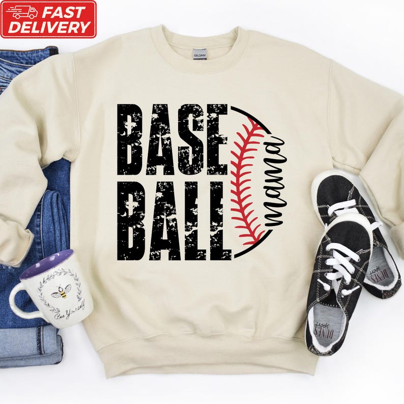 A beige sweatshirt with the text "BASE BALL" printed on it, along with a baseball graphic and a red baseball glove. Next to the sweatshirt are a pair of black and white sneakers, and a white mug with a floral design.