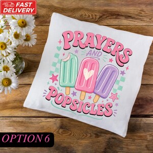 A white pillow with a colorful design featuring three popsicles and the text "Prayers and Popsicles" on it, placed on a wooden surface next to a vase of white flowers.