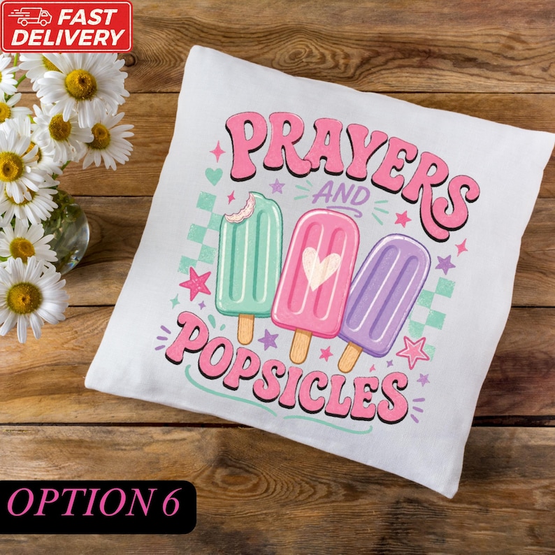 A white pillow with a colorful design featuring three popsicles and the text "Prayers and Popsicles" on it, placed on a wooden surface next to a vase of white flowers.